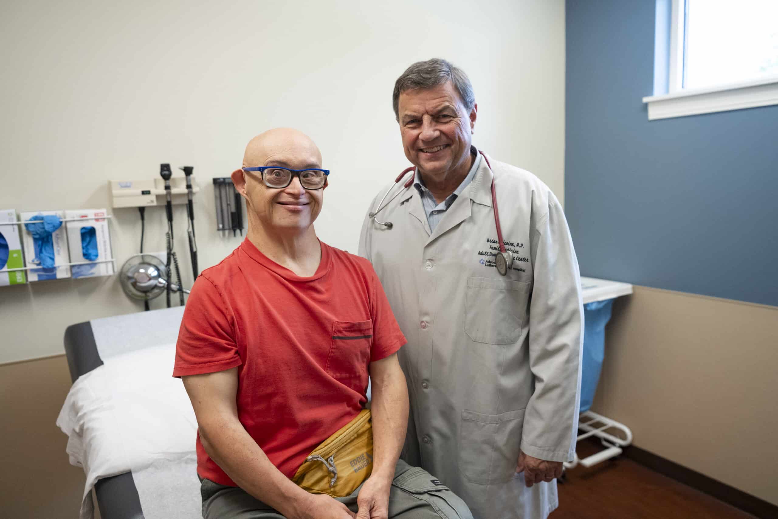 Older man with Down syndrome wearing glasses and sitting on an exam table. Male doctor in gray exam coat standing next to exam table.